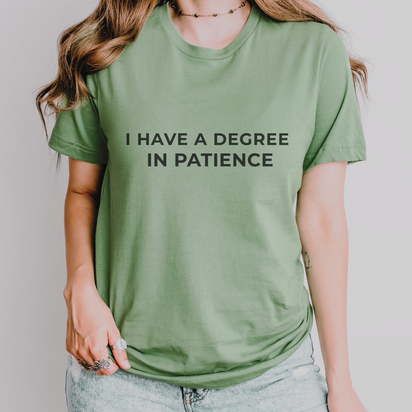 Sage green teacher t-shirt with “I Have a Degree in Patience” text, styled in a soft, neutral teacher-friendly setting.